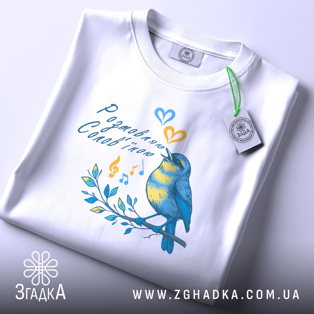 Футболка розмовляю солов’їною білого кольору, складена на столі, фон світло-сірий, принт з пташкою, що співає, в жовто-блакитних тонах, з етикеткою на зеленому шнурку.