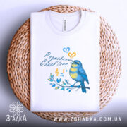 Футболка розмовляю солов’їною білого кольору, складена на плетеній основі, фон світло-сірий, принт з пташкою, що співає, в жовто-блакитних тонах, оточена нотами та листям.