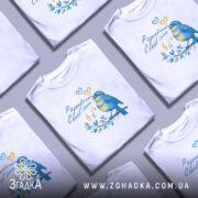 Футболка розмовляю солов’їною білого кольору, зображена на манекені з блискучою поверхнею, фон світло-сірий, принт з пташкою, що співає, в жовто-блакитних тонах, оточена нотами та листям.