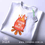 Футболка розпали моє багаття, біла, зображена зверху, складена на столі, фон світлий, принт з вогнем та текстом "РОЗПАЛИ МОЄ БАГАТТЯ" на червоному фоні, з зеленим ярликом.
