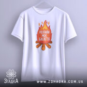 Футболка розпали моє багаття, біла, зображена з переднього ракурсу, підвішена на вішалці, фон світлий, принт з вогнем та текстом "РОЗПАЛИ МОЄ БАГАТТЯ" на червоному фоні, під ним зображення дров.