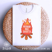 Футболка розпали моє багаття, біла, зображена зверху, складена на плетеній основі, фон світлий, принт з вогнем та текстом "РОЗПАЛИ МОЄ БАГАТТЯ" на червоному фоні, під ним зображення дров.