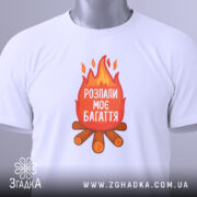 Футболка розпали моє багаття, біла, зображена зверху, складена на столі, фон світлий, принт з вогнем та текстом "РОЗПАЛИ МОЄ БАГАТТЯ" на червоному фоні, з зеленим ярликом.