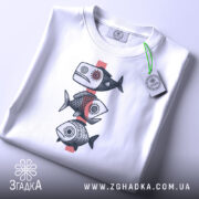 Футболка рибний етно, біла, зображена зверху на плоскій поверхні, фон світло-сірий, принт з трьох риб у стилі етно, з етикеткою зеленого кольору.