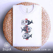 Футболка рибний етно, біла, зображена зверху на плоскій поверхні, фон світло-сірий, принт з трьох риб у стилі етно, розташована на плетеній основі.