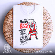 Футболка Санта новорічний вайб, біла, складена на плетеному кошику, фон світло-сірий, принт з веселим Сантою, текст 'Happy New Year!' у червоному кольорі.