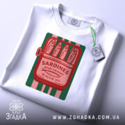 Футболка сардини білого кольору, складена на столі, з принтом у вигляді консервної банки з сардинами, червоний текст на зеленому фоні, з зеленим ярликом.