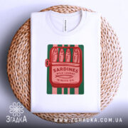Футболка сардини білого кольору, складена на плетеній кошику, з принтом у вигляді консервної банки з сардинами, червоний текст на зеленому фоні, фон білий.