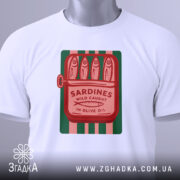 Футболка сардини білого кольору, складена на столі, з принтом у вигляді консервної банки з сардинами, червоний текст на зеленому фоні, з зеленим ярликом.