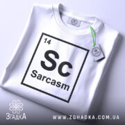 Футболка сарказм елемент: складена біла футболка з принтом 'Sc Sarcasm' у вигляді таблиці Менделєєва, видно етикетку з зеленим шнурком, фон світло-сірий, ракурс зверху.
