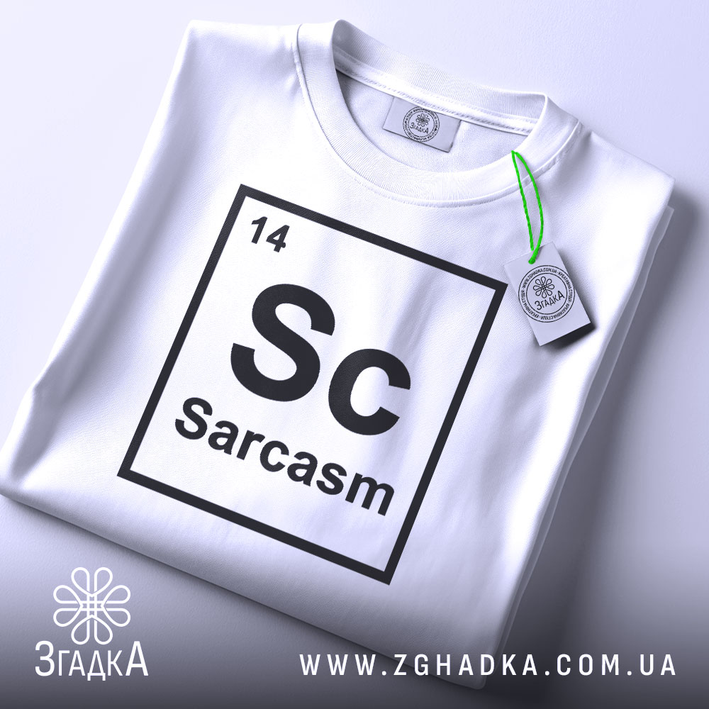Футболка сарказм елемент: складена біла футболка з принтом 'Sc Sarcasm' у вигляді таблиці Менделєєва, видно етикетку з зеленим шнурком, фон світло-сірий, ракурс зверху.
