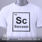 Футболка сарказм елемент: складена біла футболка з принтом 'Sc Sarcasm' у вигляді таблиці Менделєєва, видно етикетку з зеленим шнурком, фон світло-сірий, ракурс зверху.