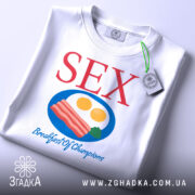 Футболка секс сніданок, біла, зображена з верхнього ракурсу, складена на поверхні, фон світлий, принт містить червоний текст 'SEX' та кольорову ілюстрацію сніданку з яєць та бекону, з етикеткою на рукаві.