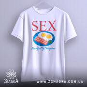 Футболка секс сніданок, біла, зображена на вішалці, фон нейтральний, принт містить червоний текст 'SEX' та кольорову ілюстрацію сніданку з яєць та бекону.