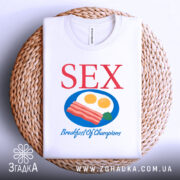 Футболка секс сніданок, біла, зображена з верхнього ракурсу, складена на плетеній основі, фон світлий, принт містить червоний текст 'SEX' та кольорову ілюстрацію сніданку з яєць та бекону.