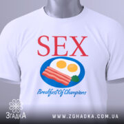 Футболка секс сніданок, біла, зображена з верхнього ракурсу, складена на поверхні, фон світлий, принт містить червоний текст 'SEX' та кольорову ілюстрацію сніданку з яєць та бекону, з етикеткою на рукаві.