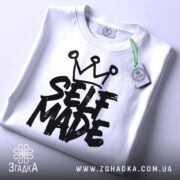 Футболка self made білого кольору, складена на плоскій поверхні, фон світло-сірий, принт чорного кольору з написом 'SELF MADE' та короною зверху, прикріплена етикетка з зеленим шнурком.