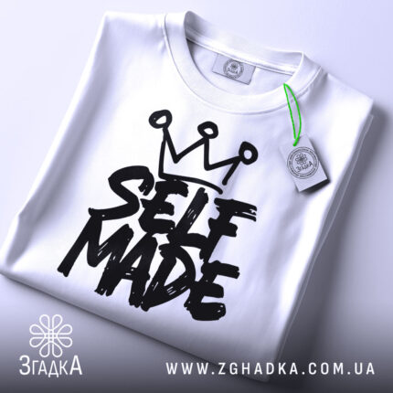 Футболка self made білого кольору, складена на плоскій поверхні, фон світло-сірий, принт чорного кольору з написом 'SELF MADE' та короною зверху, прикріплена етикетка з зеленим шнурком.