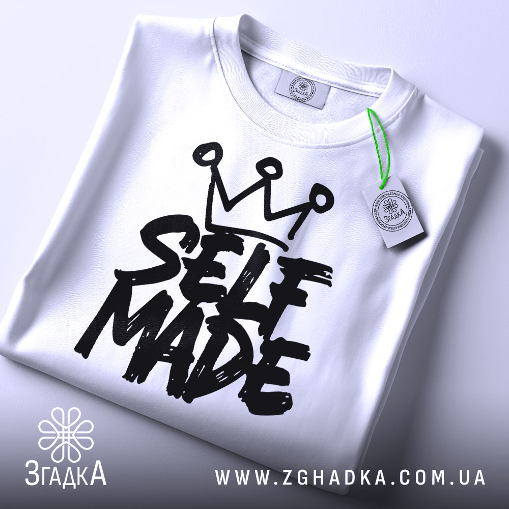Футболка self made білого кольору, складена на плоскій поверхні, фон світло-сірий, принт чорного кольору з написом 'SELF MADE' та короною зверху, прикріплена етикетка з зеленим шнурком.