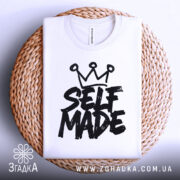 Футболка self made білого кольору, складена на плоскій поверхні з плетеним кошиком на фоні, принт чорного кольору з написом 'SELF MADE' та короною зверху.