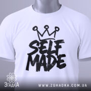 Футболка self made білого кольору, складена на плоскій поверхні, фон світло-сірий, принт чорного кольору з написом 'SELF MADE' та короною зверху, прикріплена етикетка з зеленим шнурком.