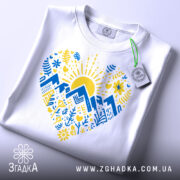 Футболка серце карпатський мотив, біла, зображена на столі, ракурс зверху, фон світло-сірий, принт з жовтим сонцем, синіми горами та рослинними візерунками, на футболці є етикетка.