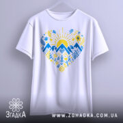 Футболка серце карпатський мотив, біла, підвішена на вішалці, фон світло-сірий, принт з жовтим сонцем, синіми горами та рослинними елементами, виглядає стильно.