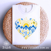 Футболка серце карпатський мотив, біла, зображена на плетеній основі, ракурс зверху, фон білий, принт з жовтим сонцем, синіми горами та рослинними візерунками.