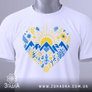 Футболка серце карпатський мотив, біла, зображена на столі, ракурс зверху, фон світло-сірий, принт з жовтим сонцем, синіми горами та рослинними візерунками, на футболці є етикетка.