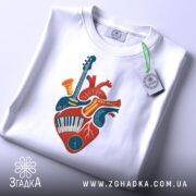 Футболка серце музиканта, біла, зображена зверху, на фоні світло-сірого кольору, принт з серцем, що містить музичні інструменти, прикрашений етикеткою з логотипом бренду.