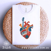 Футболка серце музиканта, біла, зображена зверху на плетеній кошику, на фоні світло-сірого кольору, принт з серцем, що містить музичні інструменти, виконаний в яскравих кольорах.