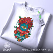 Футболка серце око мотив, складена футболка на білому фоні, ракурс зверху, принт з червоним серцем, оком та квітами, напис 'Заплющити очі, бачити серцем', етикетка з зеленим шнурком.