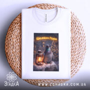 Футболка Щасливого Різдва білого кольору, складена на плетеній основі, фон світлий, принт з птахом та ліхтарем на снігу, текст "Щасливого Різдва" у верхній частині принта.