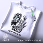 Футболка ще п’ять разів. Біла футболка, зображена зверху, на ній принт з персонажем, фон світлий, на футболці є етикетка з зеленим шнурком, деталі принта яскраві.