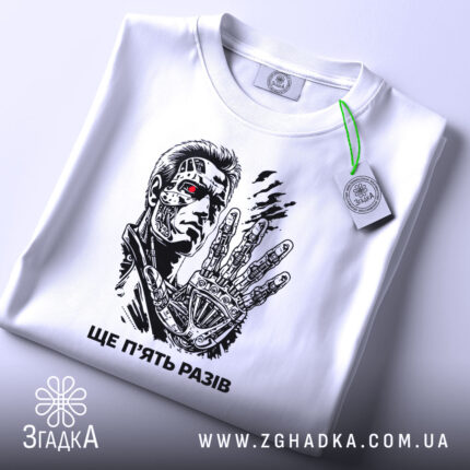 Футболка ще п’ять разів. Біла футболка, зображена зверху, на ній принт з персонажем, фон світлий, на футболці є етикетка з зеленим шнурком, деталі принта яскраві.
