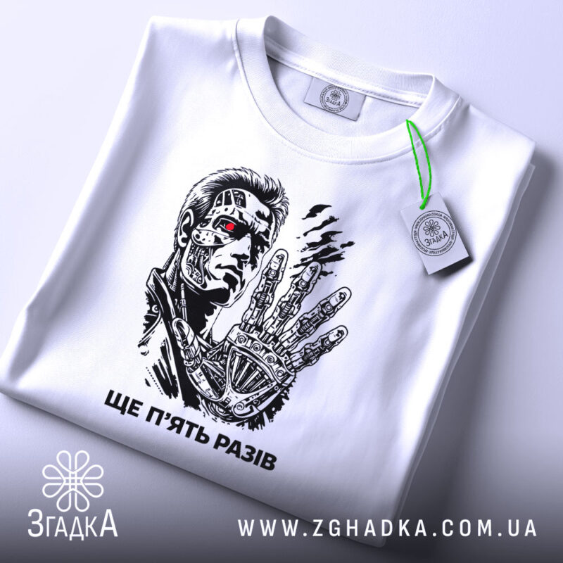 Футболка ще п’ять разів. Біла футболка, зображена зверху, на ній принт з персонажем, фон світлий, на футболці є етикетка з зеленим шнурком, деталі принта яскраві.