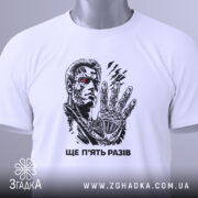 Футболка ще п’ять разів. Біла футболка, зображена зверху, на ній принт з персонажем, фон світлий, на футболці є етикетка з зеленим шнурком, деталі принта яскраві.