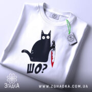 Футболка шо чорний кіт білого кольору, зображена зверху, на ній видно етикетку з зеленим шнурком, фон світло-сірий, принт з чорним котом, що тримає ніж з червоною лезом, текст 'ШО?' під котом.