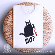 Футболка шо чорний кіт білого кольору, зображена зверху на плетеній кошику, фон білий, принт з чорним котом, що тримає ніж з червоною лезом, текст 'ШО?' під котом.