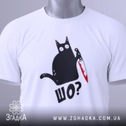 Футболка шо чорний кіт білого кольору, зображена зверху, на ній видно етикетку з зеленим шнурком, фон світло-сірий, принт з чорним котом, що тримає ніж з червоною лезом, текст 'ШО?' під котом.