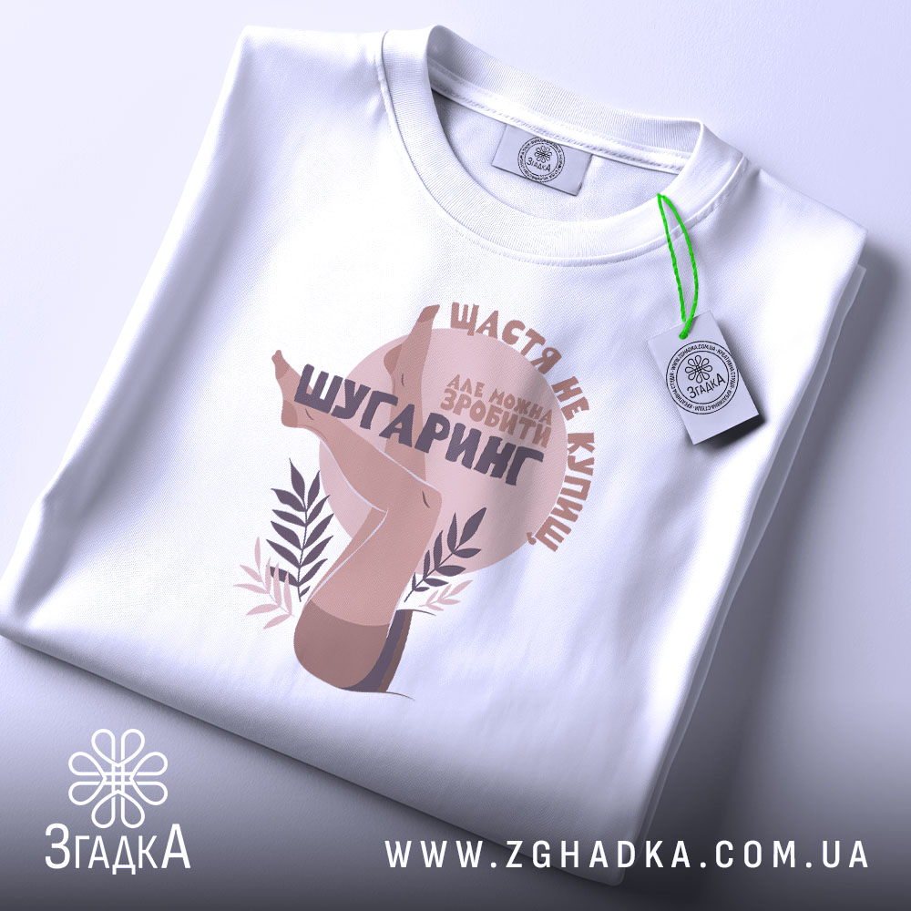 Футболка шугаринг без ілюзій, біла, складена на поверхні, фон світло-сірий, принт з зображенням жіночих ніг та текстом у коричневих тонах, з етикеткою зеленого кольору.