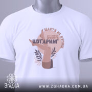 Футболка шугаринг без ілюзій, біла, складена на поверхні, фон світло-сірий, принт з зображенням жіночих ніг та текстом у коричневих тонах, з етикеткою зеленого кольору.