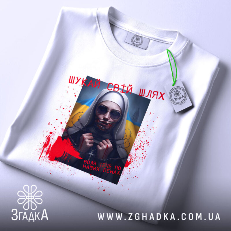 Футболка шукай свій шлях. Біла футболка, зображена зверху, з принтом на грудях, що показує жінку в темних окулярах. На фоні видно жовто-блакитний прапор, а також червоні плями, що нагадують кров, та напис українською мовою. На комірці видно етикетку.