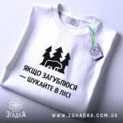 Футболка шукайте в лісі, біла, складена на поверхні, фон світло-сірий, принт чорний з ведмедем та деревами, текст українською мовою, з етикеткою зеленого кольору.
