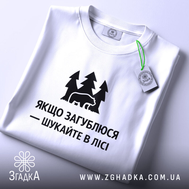 Футболка шукайте в лісі, біла, складена на поверхні, фон світло-сірий, принт чорний з ведмедем та деревами, текст українською мовою, з етикеткою зеленого кольору.