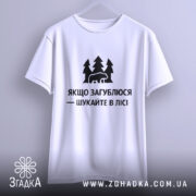 Футболка шукайте в лісі, біла, підвішена на вішалці, фон світло-сірий, принт чорний з ведмедем та деревами, текст українською мовою.
