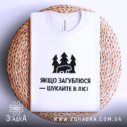 Футболка шукайте в лісі, біла, складена на плетеній кошику, фон білий, принт чорний з ведмедем та деревами, текст українською мовою.