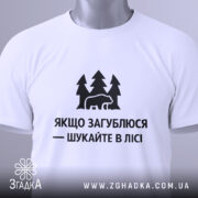 Футболка шукайте в лісі, біла, складена на поверхні, фон світло-сірий, принт чорний з ведмедем та деревами, текст українською мовою, з етикеткою зеленого кольору.