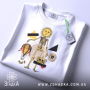 Футболка сюрреал арт скелет, біла, складена на столі, фон світло-сірий, принт з жовтим сонцем, скелетом, повітряною кулею, короною та геометричними фігурами, на футболці є етикетка з зеленим шнурком.