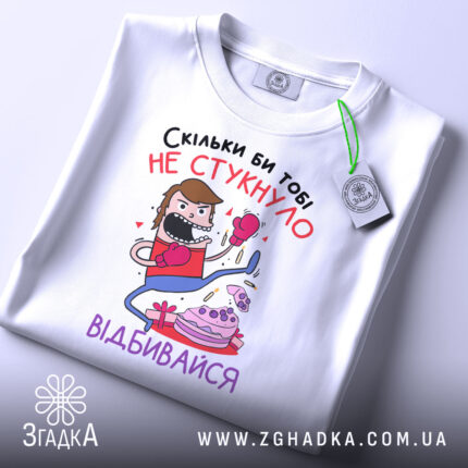 Футболка скільки би не стукнуло, білого кольору, зображена зверху, на ній видно етикетку, фон світло-сірий, принт з персонажем у боксерських рукавичках, який б'є по тортові, текст на українській мові.